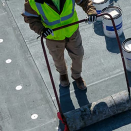 A roofer in a safety vest wheeling a roll of membrane across a flat commercial roof deck, with pails of adhesive staged beside it