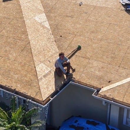 A First Coast Roofing crew member on a storm-damaged roof stripped to the decking, with the house below protected by tarps